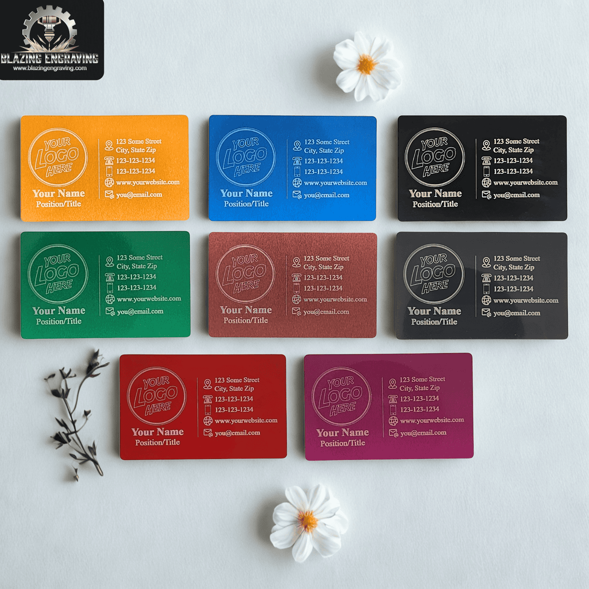 Assorted colorful laser-engraved metal business cards arranged on a light background with white flowers, each card featuring customizable logo, name, and contact details.