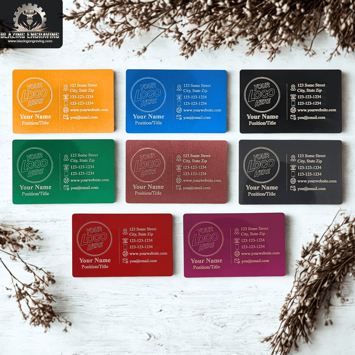 Collection of colorful laser-engraved metal business cards with customizable logo and contact details, arranged on a white wood background with dried floral accents.
