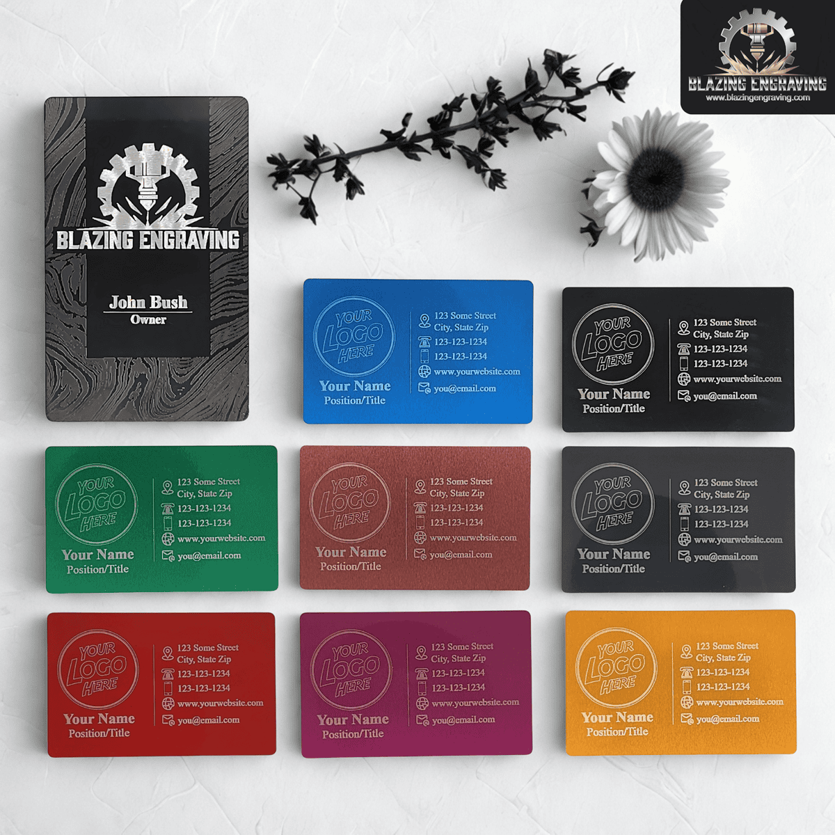 Custom engraved metal business cards in various colors and finishes, displayed with a large engraved sample card and floral accents on a white background.