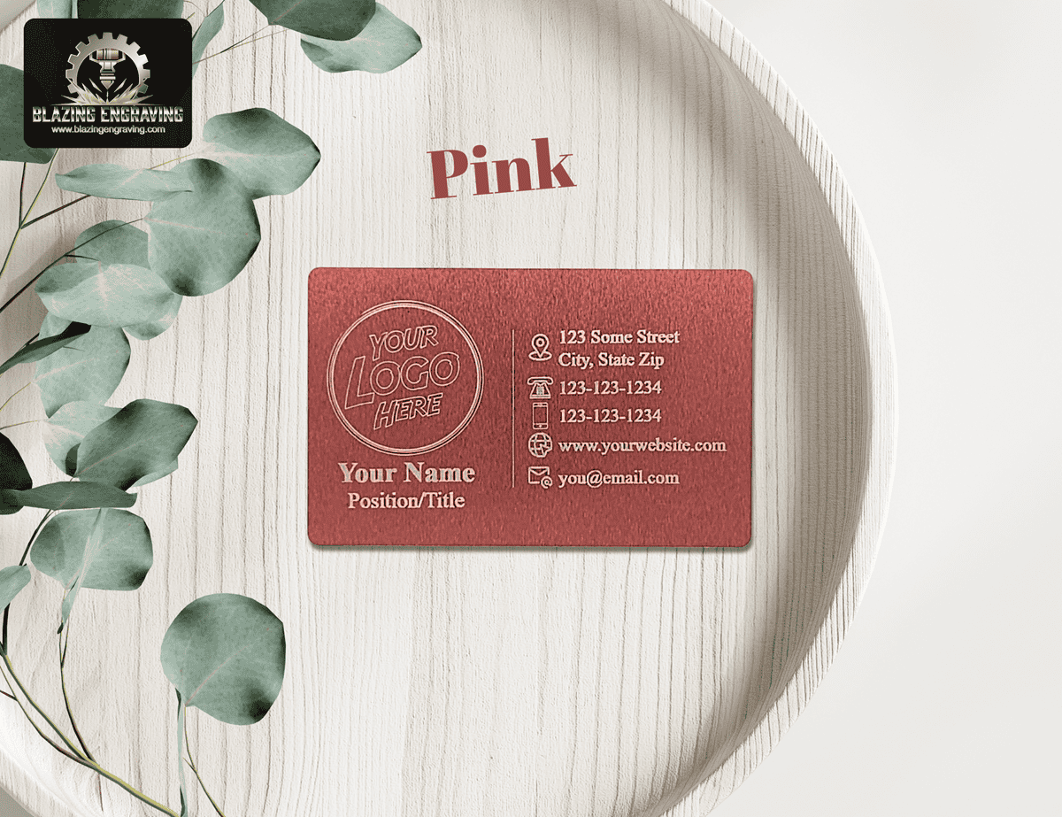 Custom laser-engraved metal business card in pink finish with space for logo, name, and contact details, displayed on a round wooden tray with eucalyptus leaves.