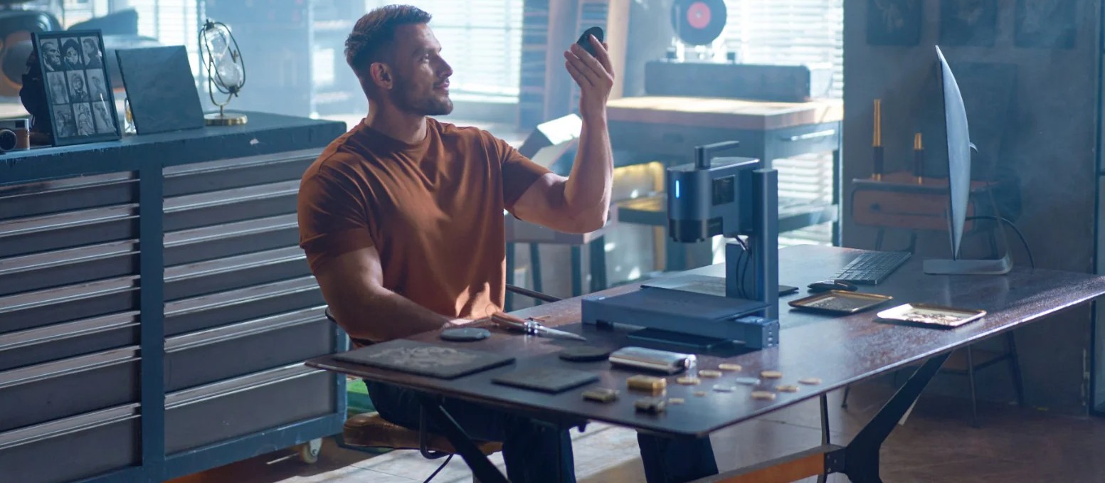 Man sitting at a workbench in a workshop, holding a small object and smiling, with a laser engraving machine and engraved items on the table, surrounded by tools and equipment in a sunlit workspace.