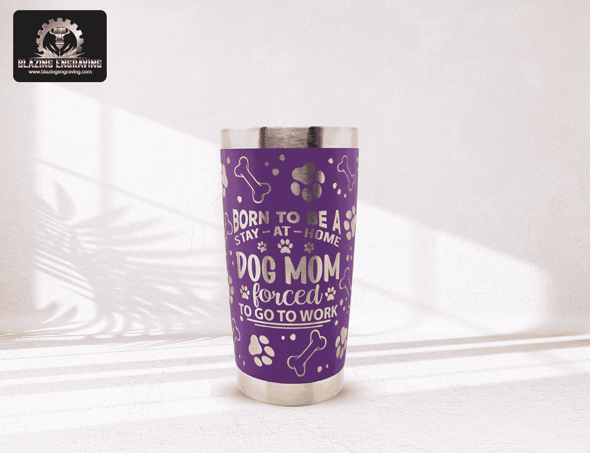 Purple stainless steel tumbler with silver laser-engraved dog paw prints, bones, and the phrase ‘Born to be a stay-at-home dog mom forced to go to work.’ Blazing Engraving logo in the top left corner.