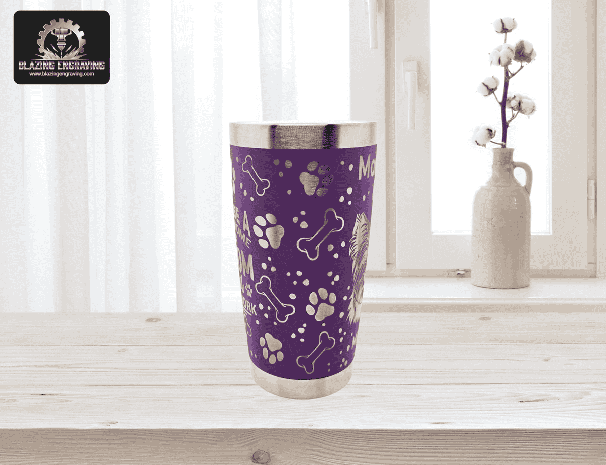 Purple stainless steel tumbler with silver laser-engraved dog paw prints, bones, and part of a dog illustration, set on a light wood surface in front of a window with sheer curtains and a vase of cotton stems. Blazing Engraving logo in the top left corner.