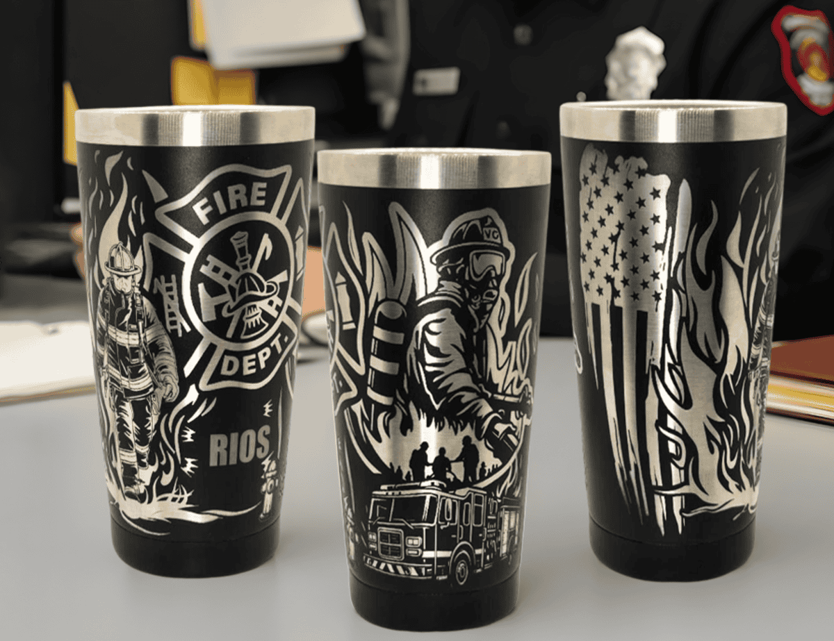 Three black stainless steel tumblers with silver laser-engraved firefighter-themed designs, including a firefighter, fire department emblem, fire truck, American flag, and flames. Displayed on a desk with a firefighter in uniform in the background.