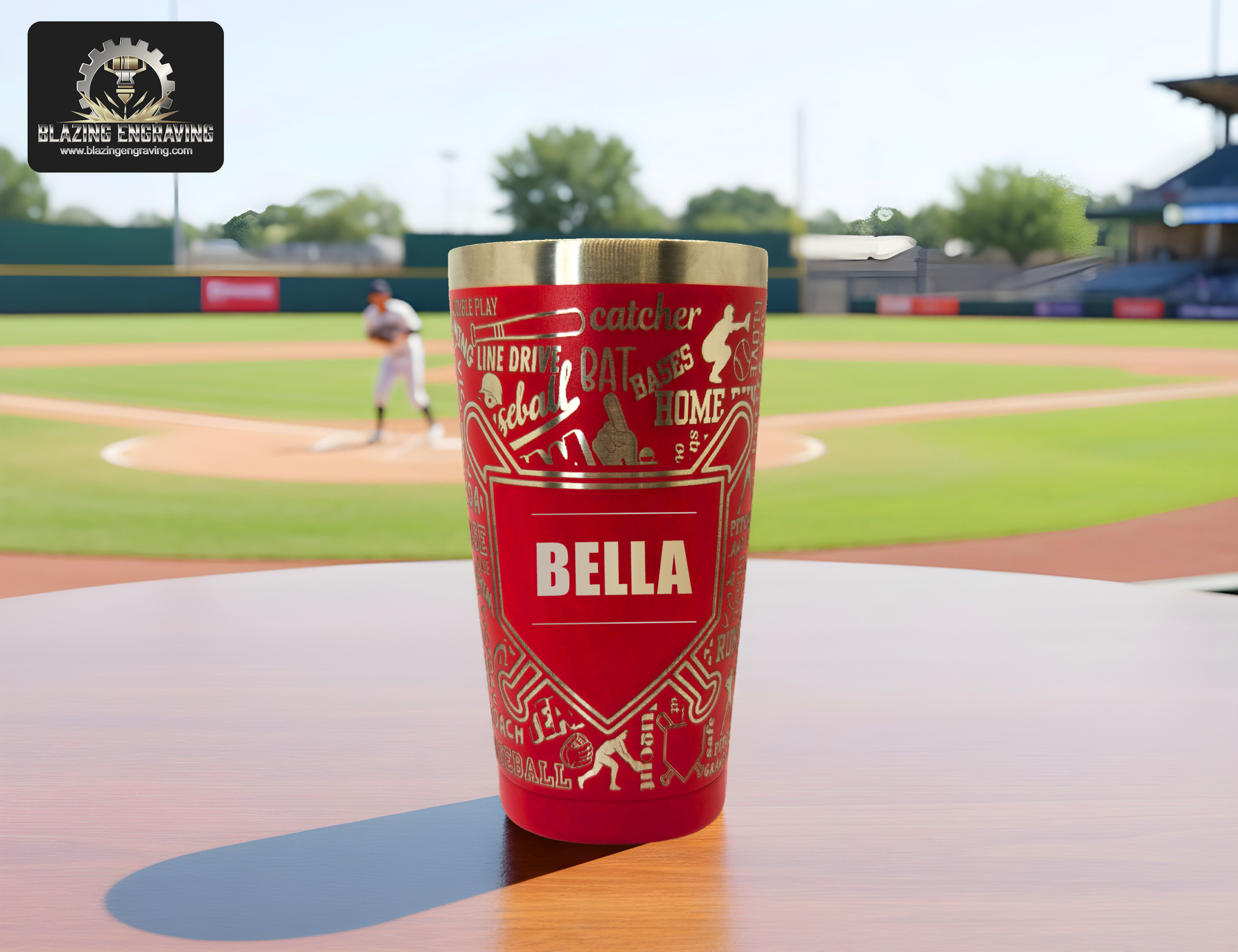 A stainless steel tumbler with a laser-engraved baseball mom design, personalized with the name "Bella," displayed on a wooden surface with a baseball field and player in the background. Blazing Engraving logo in the top left corner.
