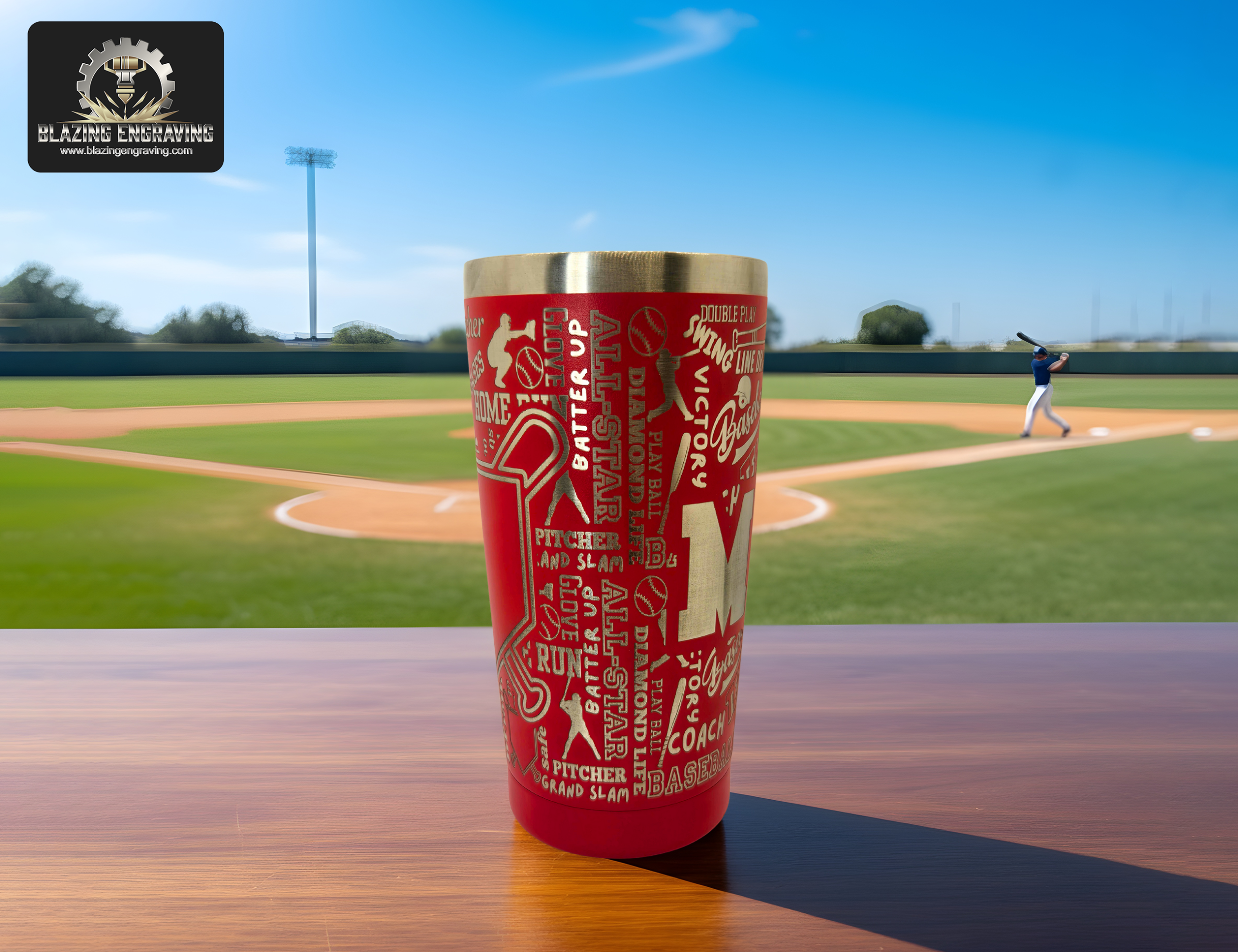 A stainless steel tumbler with a laser-engraved baseball mom design featuring baseball-themed words and player silhouettes, displayed on a wooden surface with a baseball field and batter in the background. Blazing Engraving logo in the top left corner.