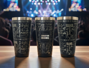 Three stainless steel tumblers showing all sides of a laser-engraved band design, personalized with the title "Director" and the name "Michel," displayed on a wooden surface with a concert stage in the background. Blazing Engraving logo in the top left corner.